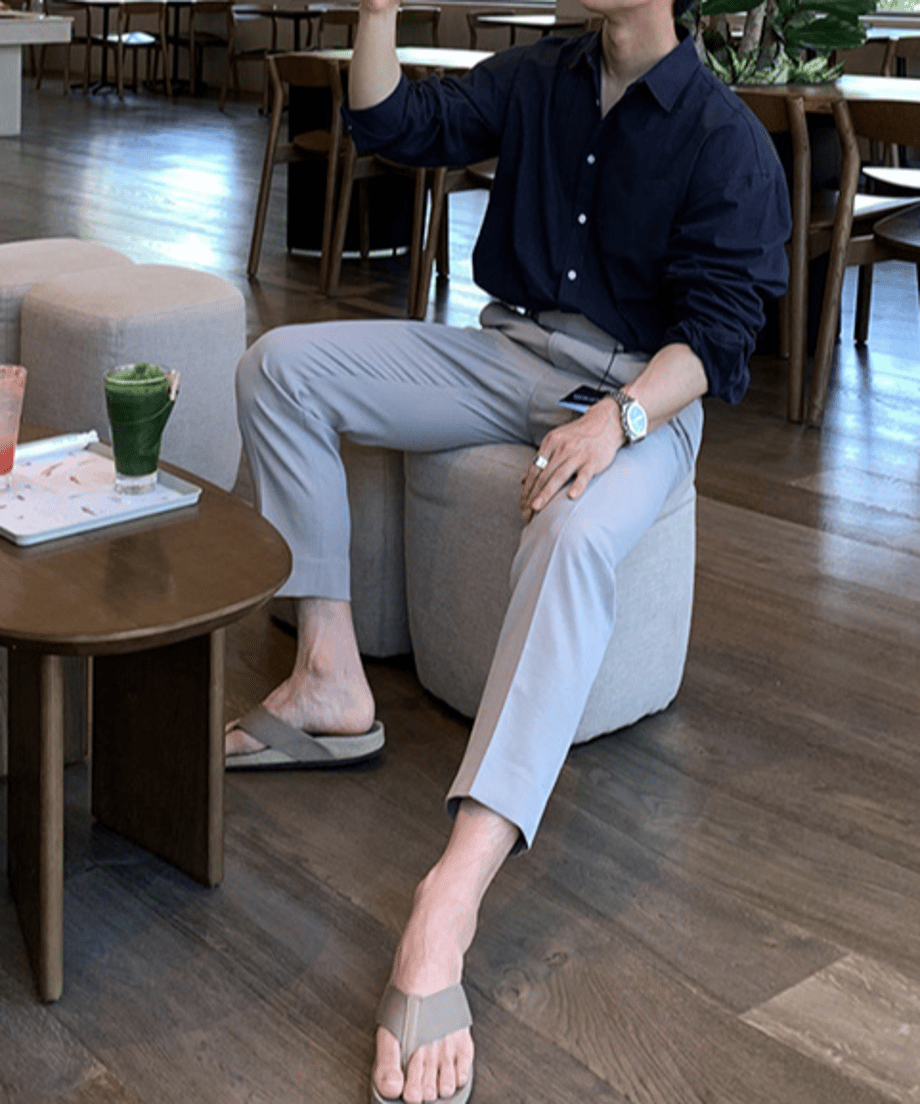 ネイビーの長袖シャツとライトグレーのスラックス、足元にベージュのトングサンダルを合わせた男性が、カフェのスツールにゆったりと腰掛けている全身コーディネートカット。
