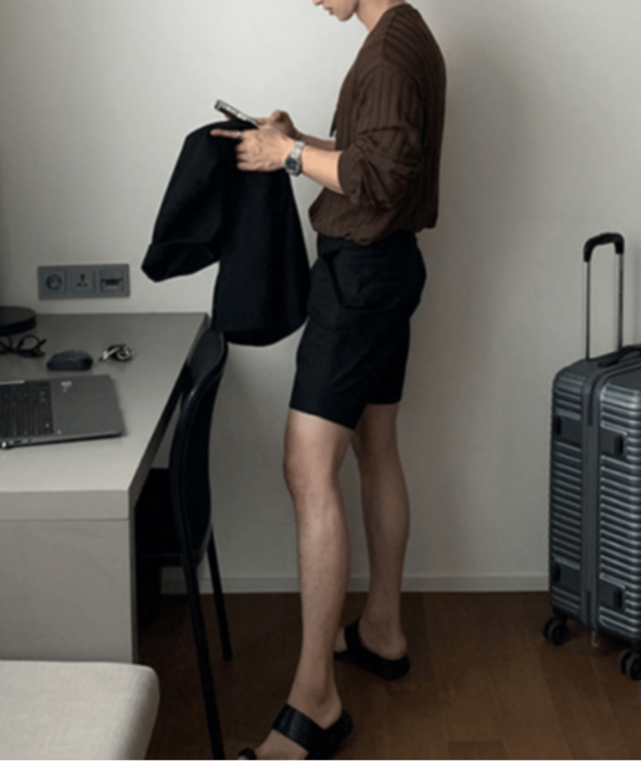 ホテルのような白い壁の部屋で、ブラウンのカーディガンに黒のハーフパンツ、足元に黒のレザートングサンダルを合わせた男性の全身サイドカット。