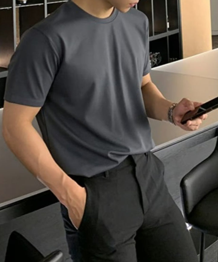 ライトグレーのクルーネックTシャツをブラックのボトムスにタックインし、スマートフォンを手に持つ男性の斜め前からのカット。