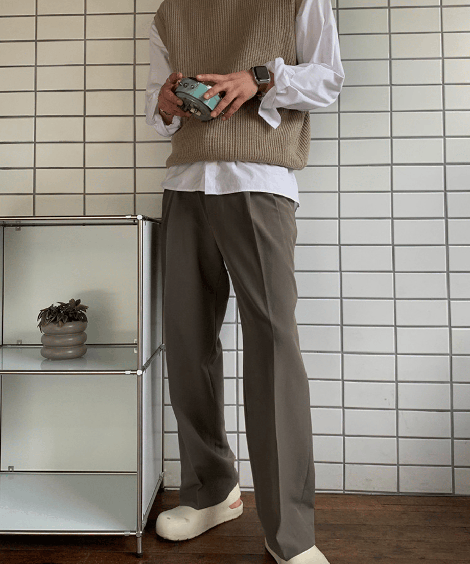 白いタイル壁の前で、ベージュのニットベストに白の長袖シャツ、ダークブラウンのワイドスラックスを合わせた男性の立ち姿。
