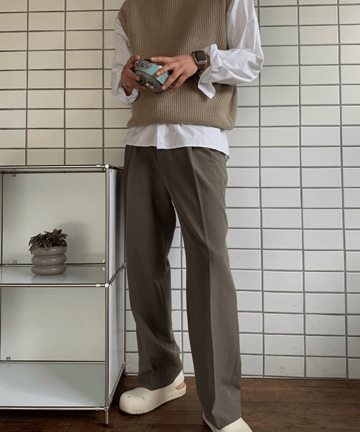 白いタイル壁の前で、ベージュのニットベストに白の長袖シャツ、ダークブラウンのワイドスラックスを合わせた男性の立ち姿。