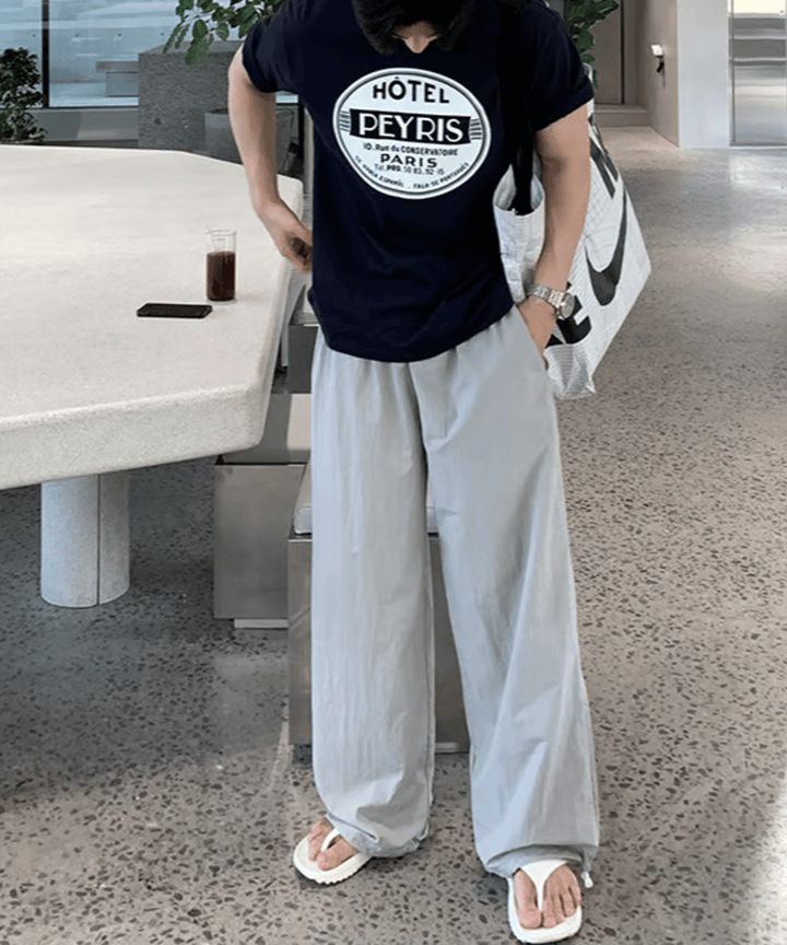 ネイビーのサークルロゴTシャツとライトグレーのイージーワイドパンツを着用し、白のトングサンダルを履いた男性の全身フロントカット。
