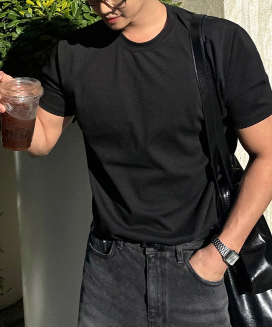 黒い半袖クルーネックTシャツにグレーのデニムパンツ、黒いレザーバッグを肩にかけ、ドリンクを手にする男性の斜め前からのアップショット。