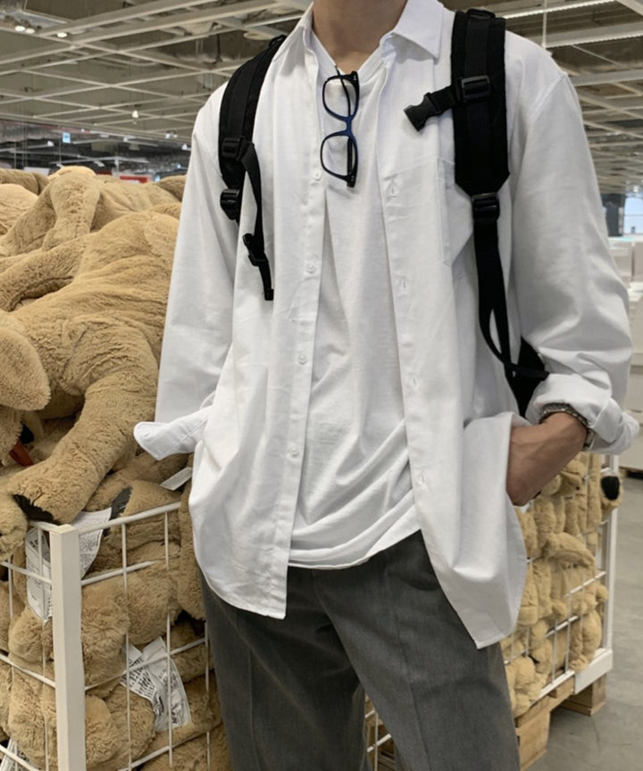胸元にメガネを掛けた白い長袖シャツにグレーのパンツを合わせ、黒いリュックサックを背負ってポケットに手を入れる男性の正面アップ。