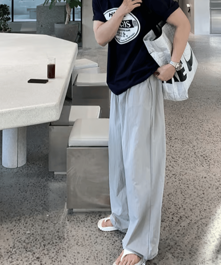 ネイビーのサークルロゴTシャツにライトグレーのルーズなワイドパンツ、足元に白のトングサンダルを合わせた男性が、大きな白いNIKEのロゴ入りトートバッグを肩に掛けて歩き出す瞬間のサイド・フロントカット。