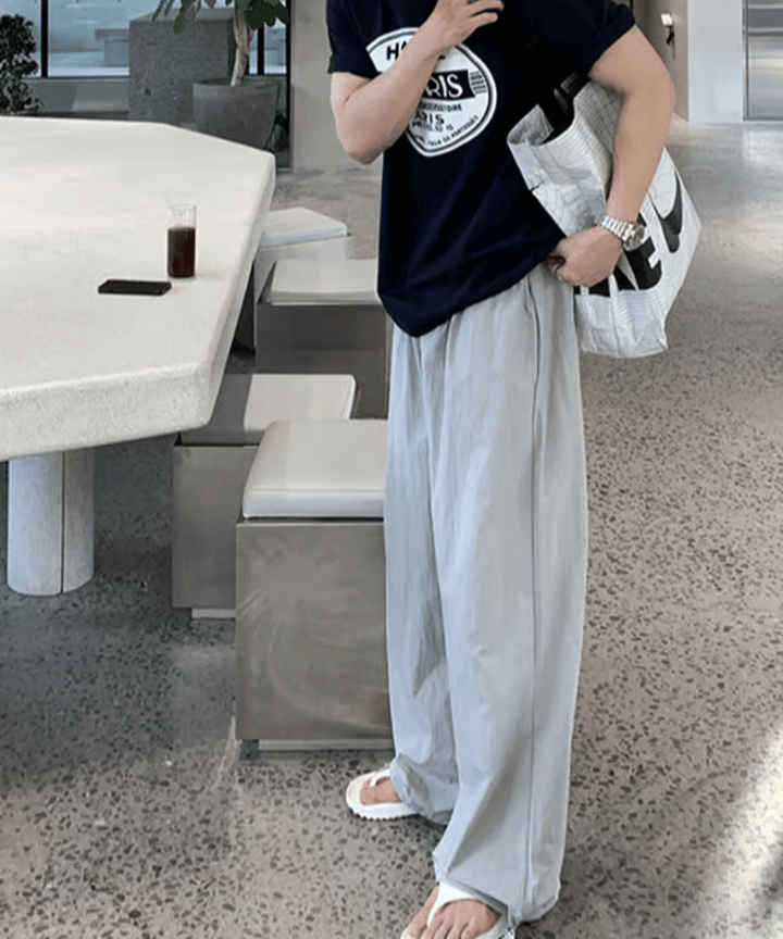 ネイビーのサークルロゴTシャツにライトグレーのルーズなワイドパンツ、足元に白のトングサンダルを合わせた男性が、大きな白いNIKEのロゴ入りトートバッグを肩に掛けて歩き出す瞬間のサイド・フロントカット。