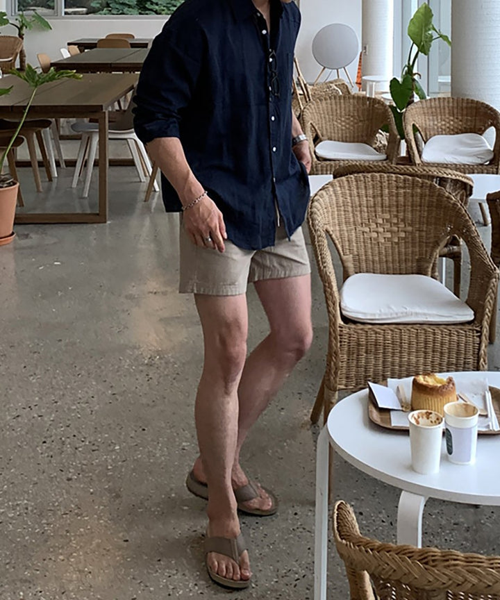 明るいカフェの店内で、ネイビーブルーの長袖リネンシャツとベージュのハーフパンツ、ブラウンのトングサンダルを着用して立つ男性の全身カット。