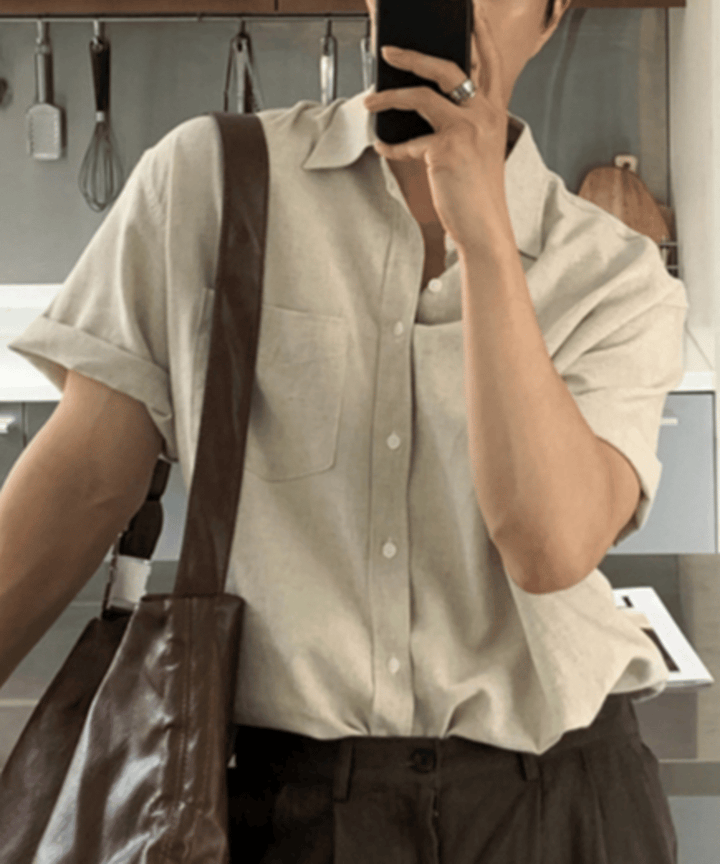 ベージュのリネン半袖シャツの袖をロールアップし、鏡越しにスマートフォンで自撮りをする男性の上半身。