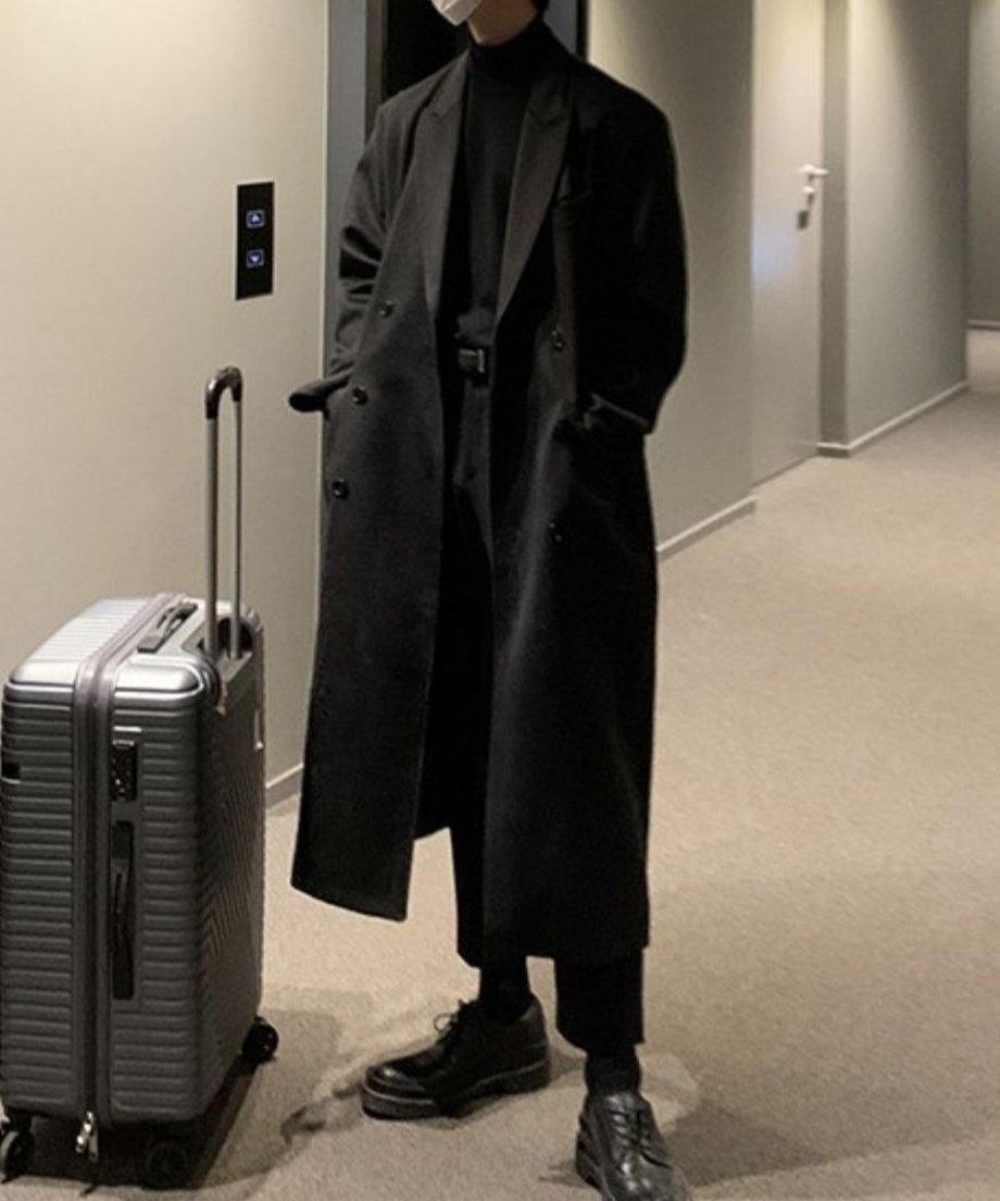 ブラックのロングチェスターコートに黒の革靴を合わせ、キャリーケースを持って立つ男性の全身カット。