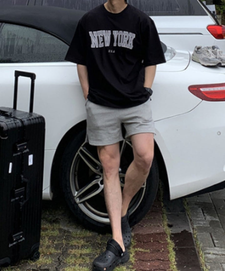 白い車の横で黒のロゴTシャツにグレーのハーフパンツ、黒のサンダルを履いて立つ男性の全身カット