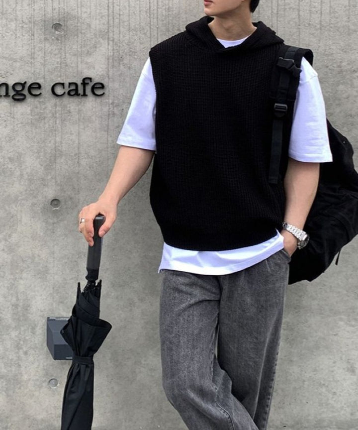 コンクリート壁を背景に、白Tシャツとブラックのフード付きニットベスト、グレーのデニムパンツを着用し、黒い傘を手にして立つ男性の正面ショット