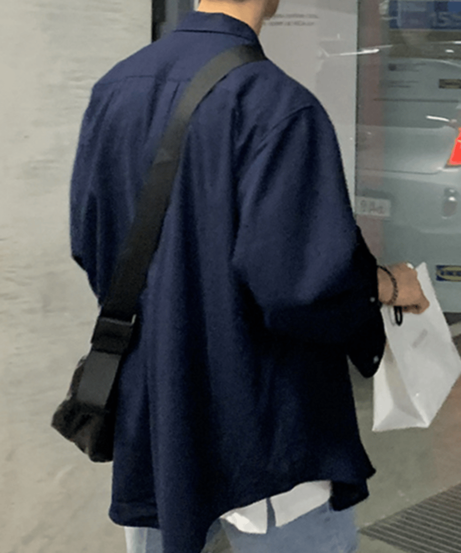 黒いショルダーバッグを斜め掛けし、ネイビーの長袖シャツの下から白いインナーを覗かせて歩く男性の後ろ姿のクローズアップ。