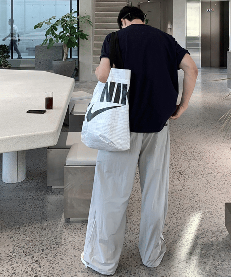 ネイビーのサークルロゴTシャツにライトグレーのワイドパンツを合わせ、NIKEの白い大きなトートバッグを肩に掛けた男性の後ろ姿（バックカット）。