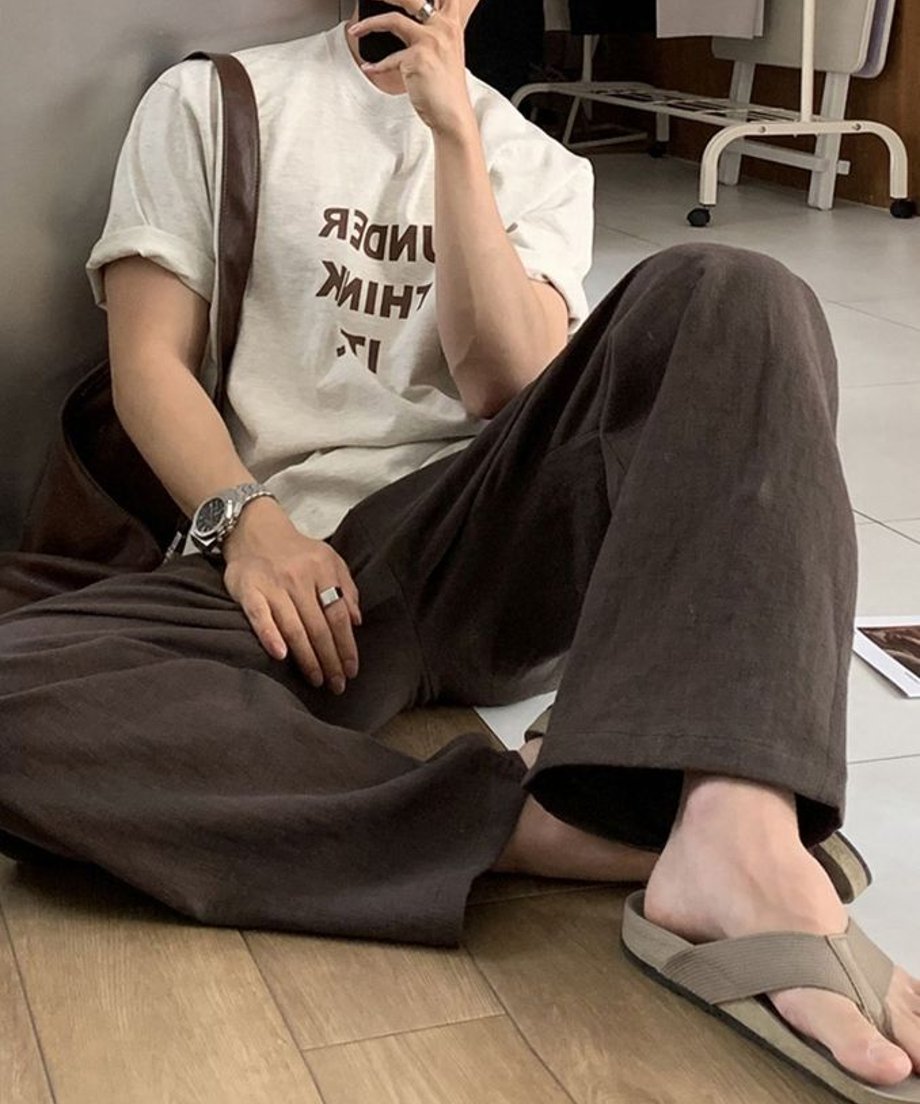 ロゴ入りのTシャツとコーヒー色のワイドパンツを着用し、床に座り込んでスマートフォンを持つ男性のカット。