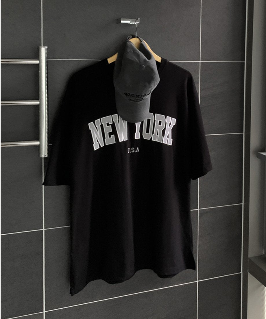 黒いタイル壁のハンガーに掛けられたブラックのNEW YORKロゴTシャツとグレーのキャップ