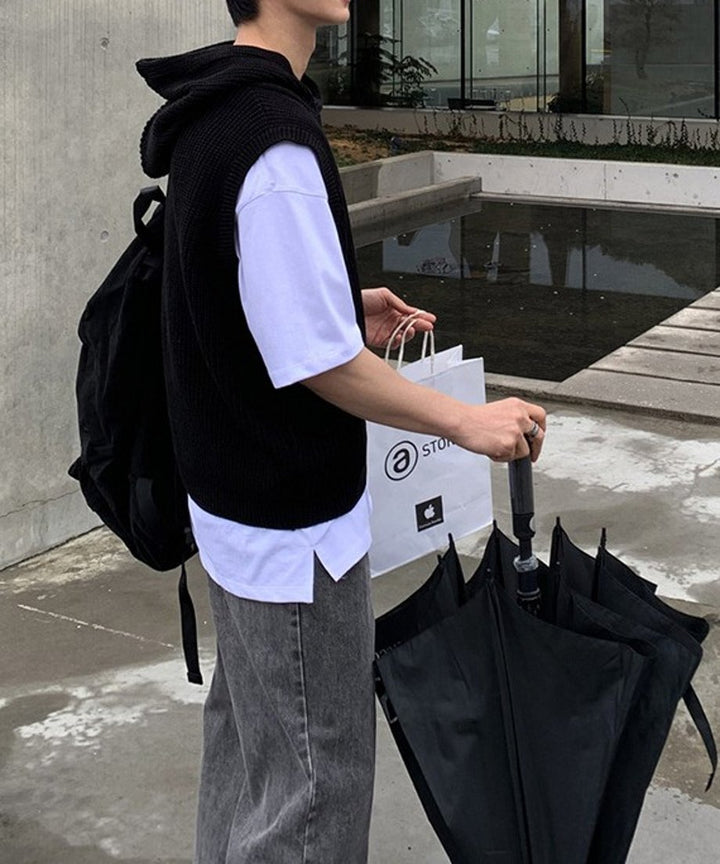 屋外の水辺近くで、ブラックのフード付きベストにリュックを背負い、白いショップバッグと大きな黒い傘を開きながら持つ男性のサイドショット