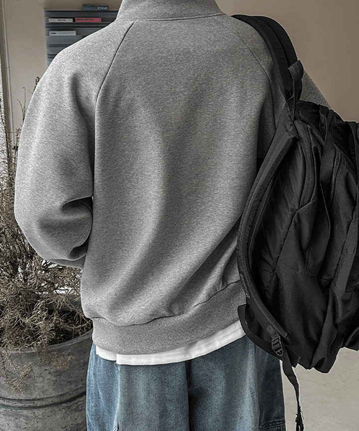 グレーのスタンドカラージップアップジャケットとデニムパンツを着用し、黒いリュックサックを背負った男性のバックスタイル