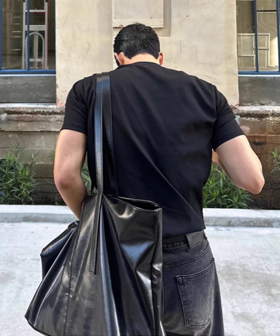 黒い半袖クルーネックTシャツに黒いデニムパンツを合わせ、黒い大きなレザーバッグを肩にかけて歩く男性の後ろ姿。