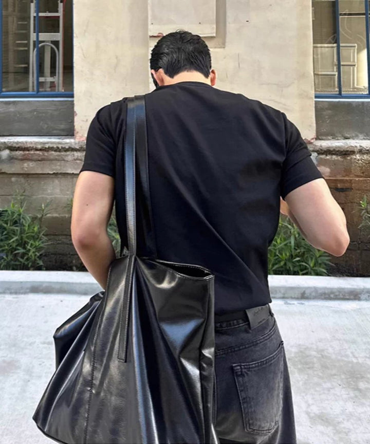 黒い半袖クルーネックTシャツに黒いデニムパンツを合わせ、黒い大きなレザーバッグを肩にかけて歩く男性の後ろ姿。