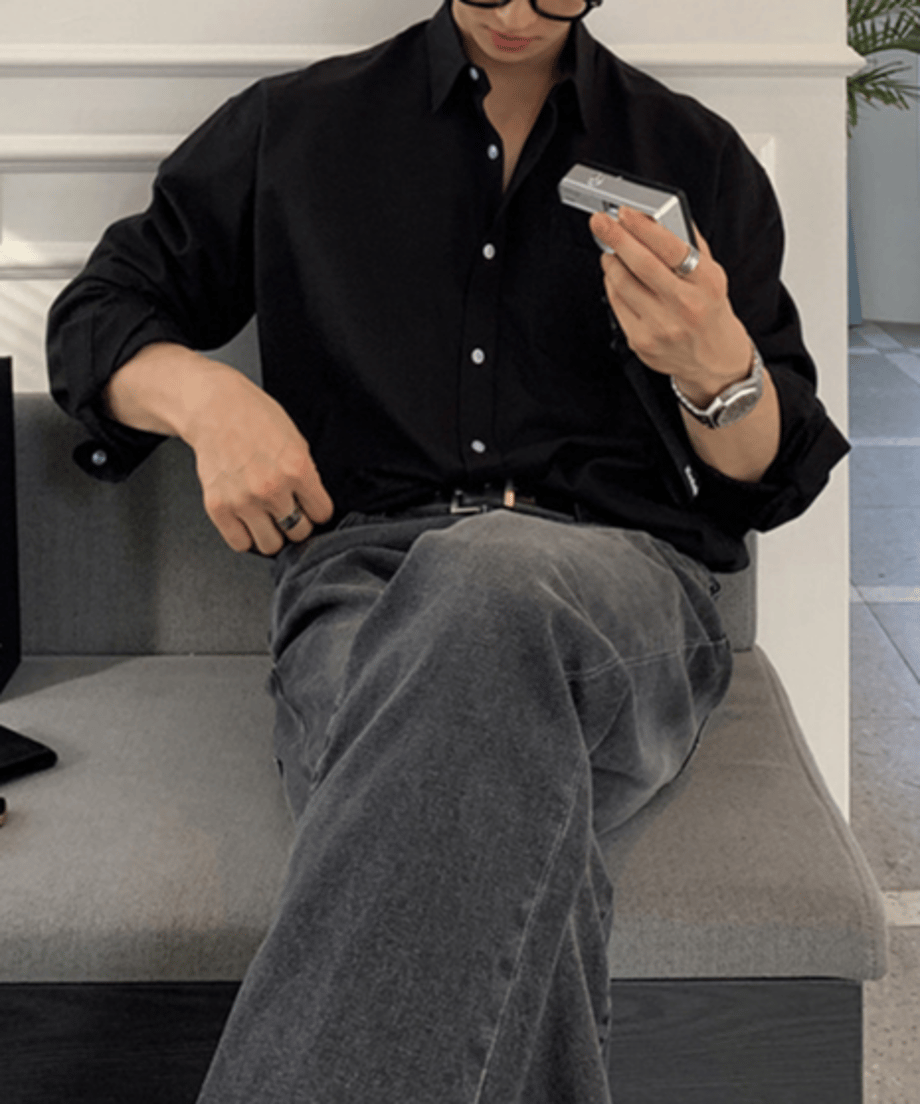 ブラックのオーバーサイズシャツを着用し、左手にシルバーのカメラを持ってソファに座る男性のチェストアップ。