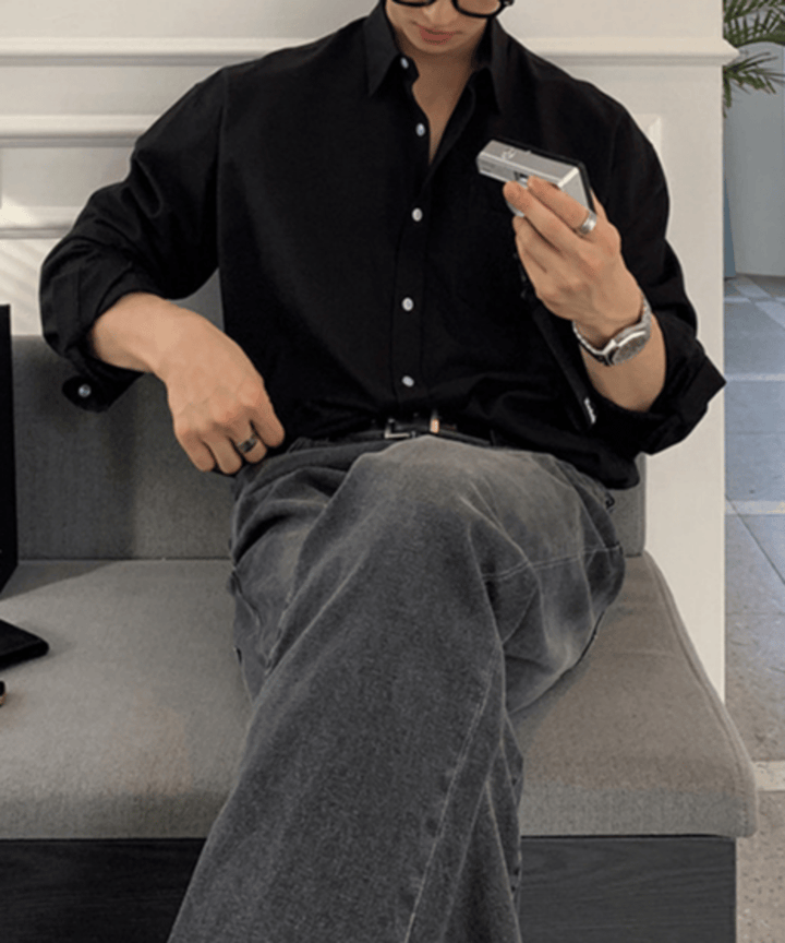 ブラックのオーバーサイズシャツを着用し、左手にシルバーのカメラを持ってソファに座る男性のチェストアップ。