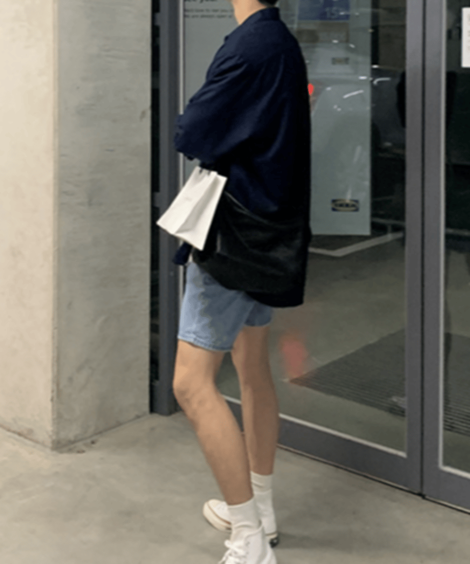 ネイビーの長袖シャツにデニムショートパンツ、白いスニーカーを合わせ、黒いショルダーバッグと白いショップバッグを持って佇む男性の横向きカット。