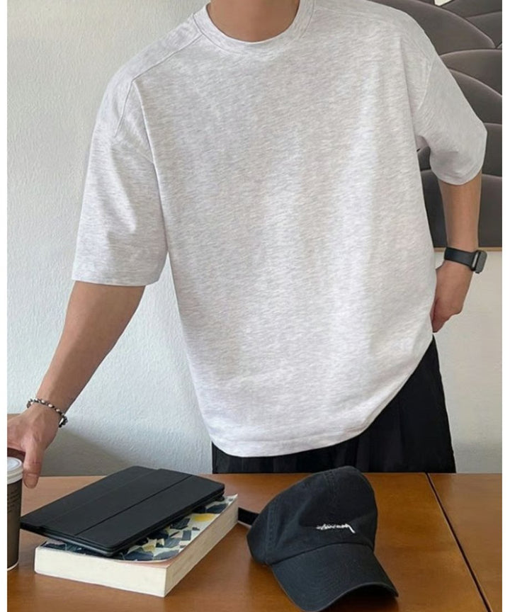 フラワーグレーのルーズTシャツにブラックのボトムスを合わせ、テーブルの上の本や黒いキャップの横に手を置いて立つ男性の斜めからのスタイリング。