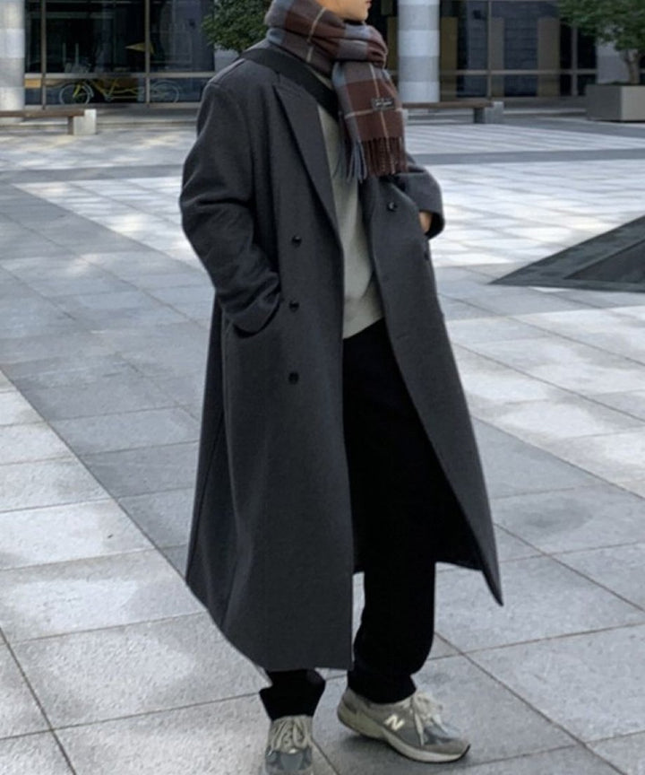 ダークグレーのロングチェスターコートのポケットに手を入れ、グレーのスニーカーを履いて歩く男性のカット。