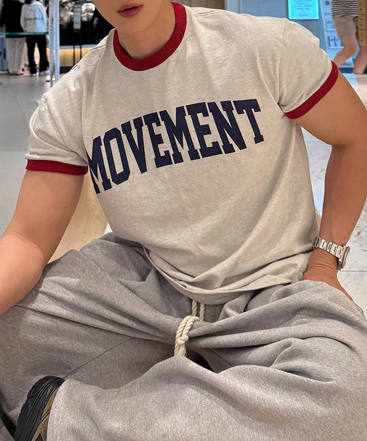 オートミール地のリンガーTシャツとグレーのスウェットパンツを着用し、床にリラックスして座る男性の斜め前からのカット。