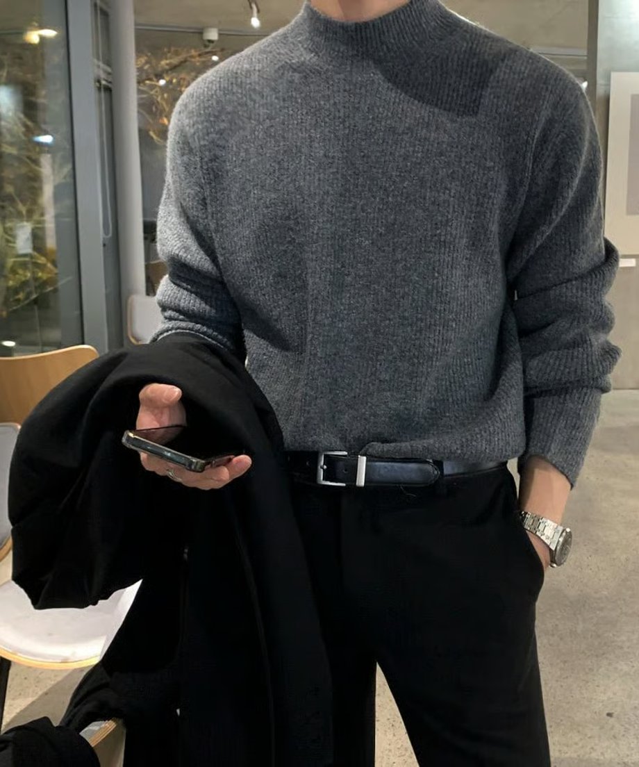 グレーのモックネックリブニットに黒のベルトとスラックスを合わせ、アウターとスマホを片手に持つ男性のスタイリッシュなフロントカット。