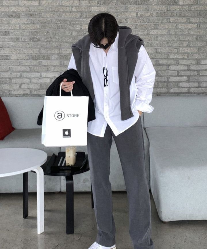 ホワイトのシャツにグレーのニットを肩掛けし、ダークグレーのパンツを穿いた男性が、白いショップバッグと黒いアウターを手に持って立っている正面全身スタイリング。