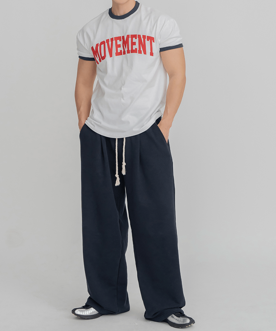 ホワイト地にネイビーのリブ、赤のロゴが入ったリンガーTシャツに、ネイビーのワイドパンツを合わせて両手をポケットに入れ、正面を見つめる男性の全身スタイリング写真。