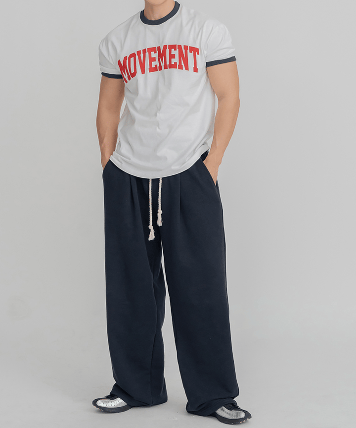 ホワイト地にネイビーのリブ、赤のロゴが入ったリンガーTシャツに、ネイビーのワイドパンツを合わせて両手をポケットに入れ、正面を見つめる男性の全身スタイリング写真。