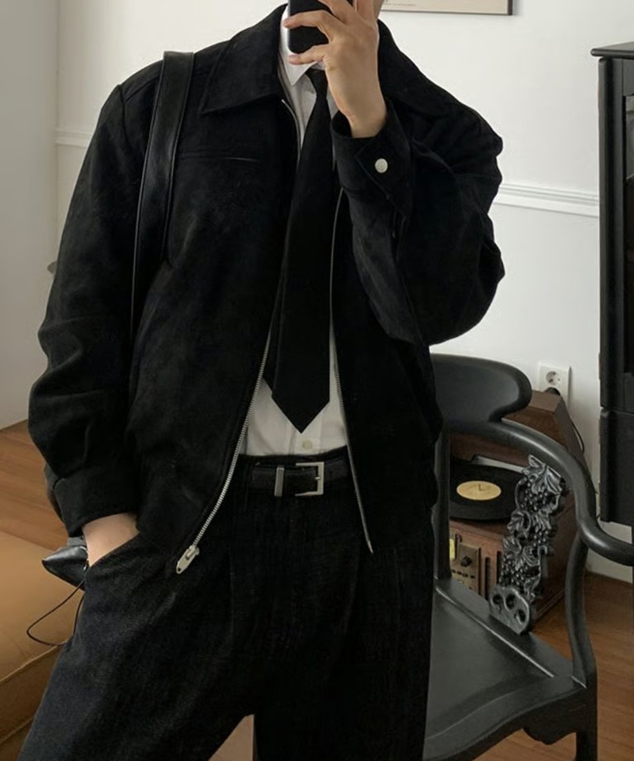 黒い椅子の横で、ブラックのフェイクスエードジップアップブルゾンのポケットに右手を入れ、白シャツとタイドアップスタイルでスマートに立つ男性のアップ
