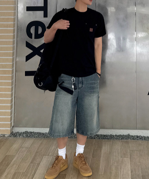 黒のTシャツにブルーのデニムショーツ、茶色のスニーカーを合わせ、黒いリュックを肩にかけた男性の正面全身コーディネート写真。