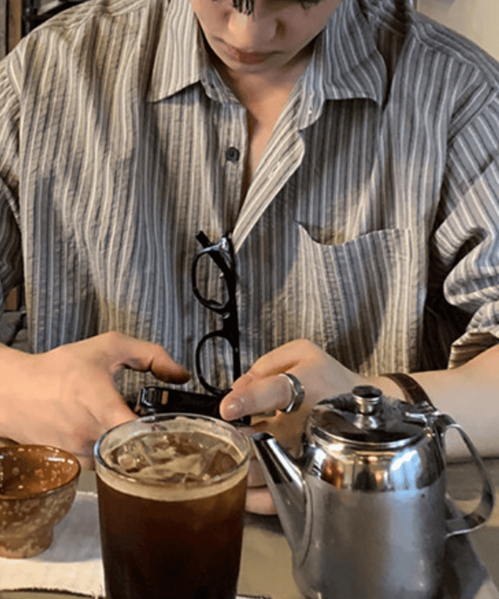 落ち着いた照明の店内で、グレーのストライプシャツを着用し、スマートフォンを操作する男性の手元。手前にはアイスコーヒーとステンレス製のティーポットが置かれています。