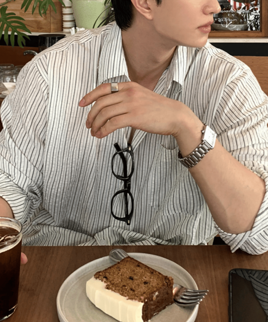 明るいカフェの店内で、胸ポケット付きのホワイトストライプ柄リラックスシャツを着用した男性のアングル。第2ボタンまで開けられた襟元には、黒いフレームのメガネが掛けられています。