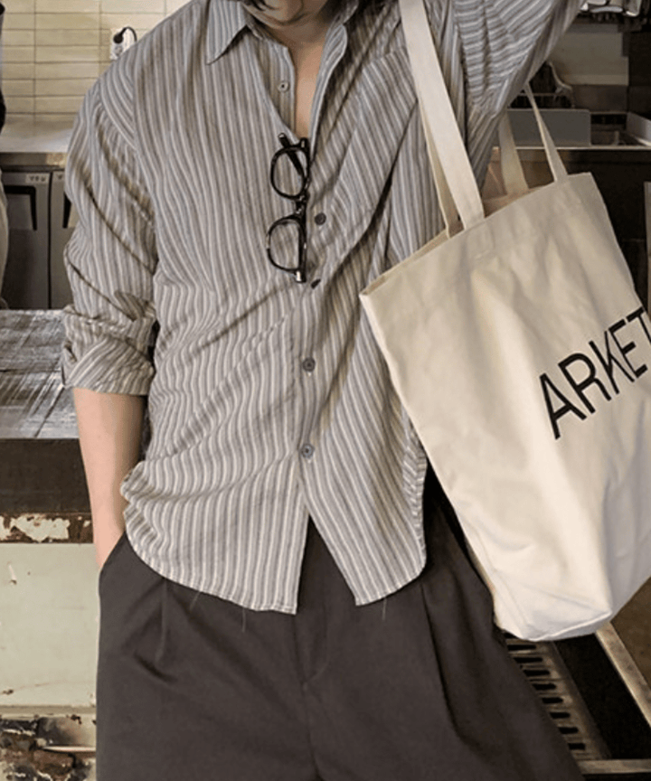 グレーのストライプ柄長袖シャツを着用し、ブラウンのワイドパンツに手を入れ、肩から「ARKET」と書かれたロゴ入りトートバッグを下げた男性の立ち姿。シャツの袖はラフにロールアップされています。