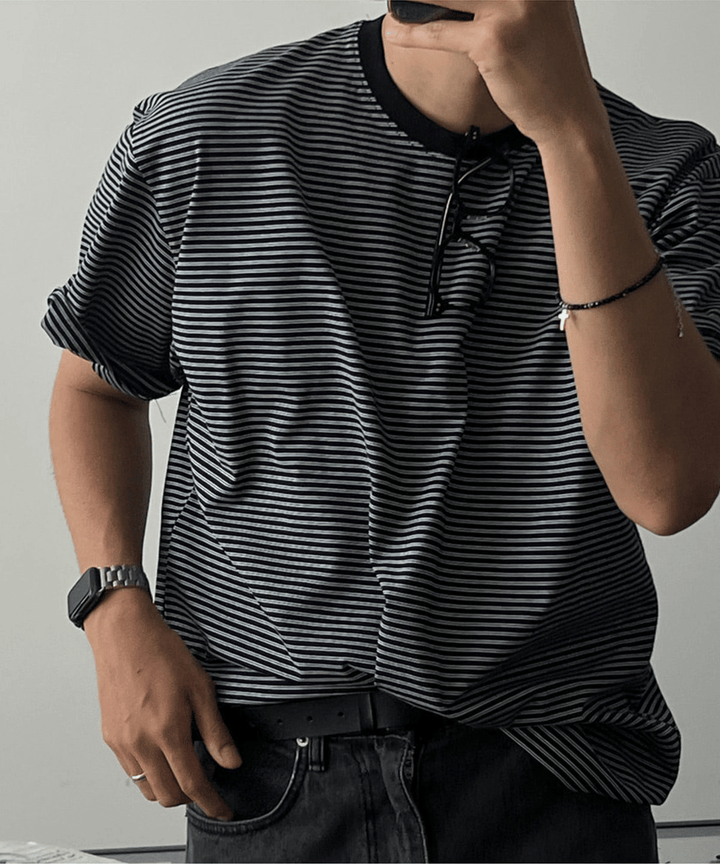 ブラックのマルチストライプ柄のTシャツを着た男性がスマホを持ちポーズを取る写真。