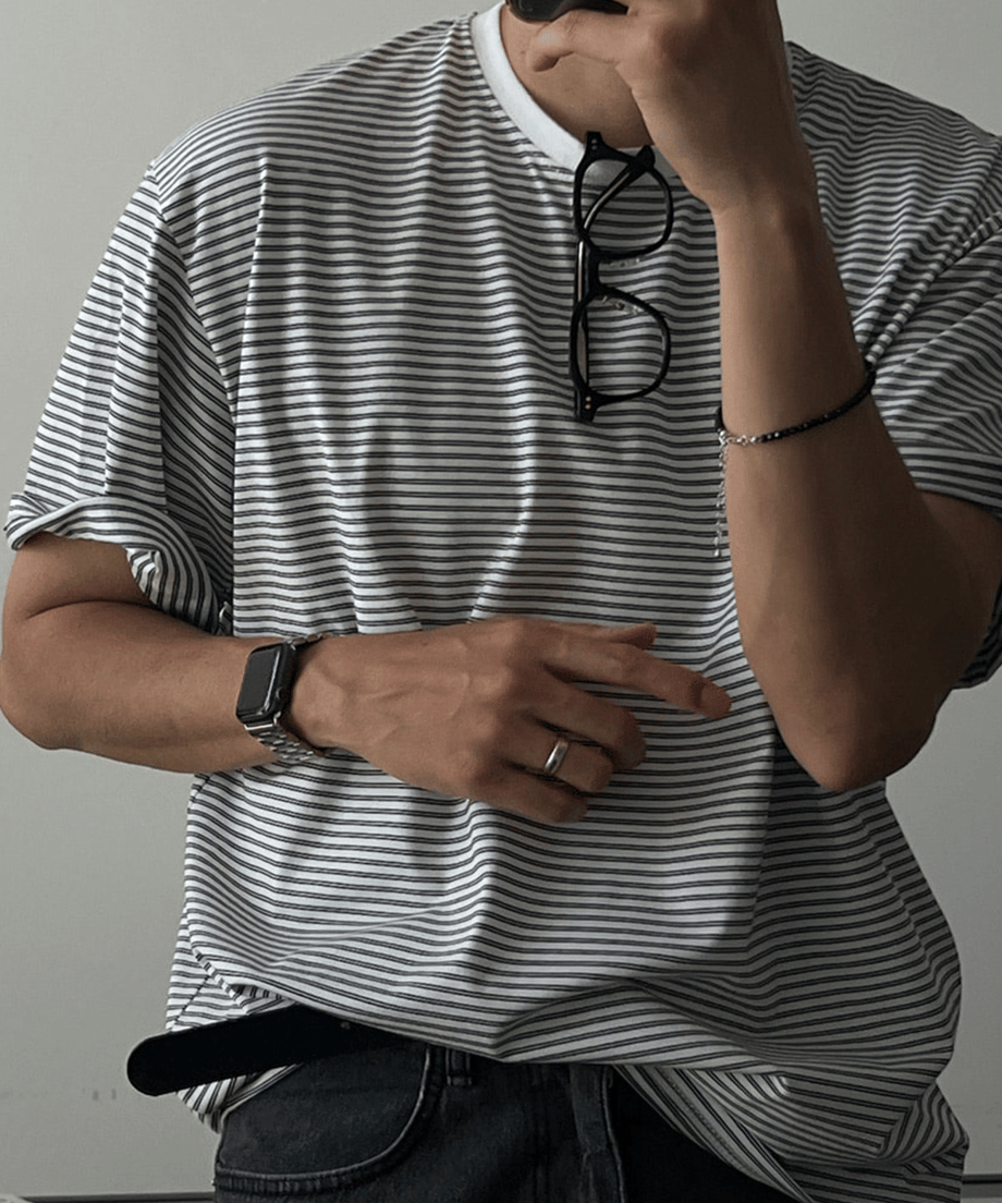 グレーのマルチストライプ柄オーバーサイズTシャツを着用した男性の正面からのアップ。首元にサングラスを掛けています。