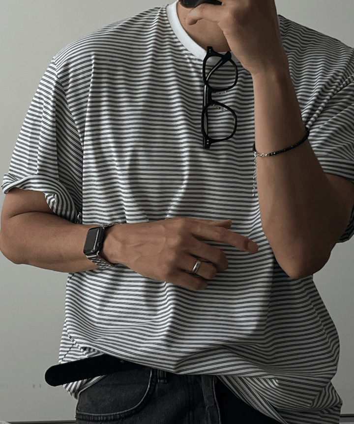 グレーのマルチストライプ柄オーバーサイズTシャツを着用した男性の正面からのアップ。首元にサングラスを掛けています。