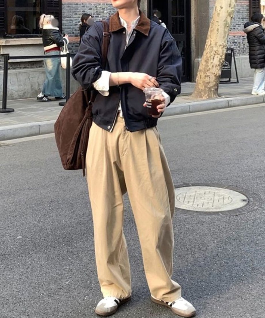 道路上でコーヒーを片手に立ち、ルーズなシルエットのダークブルーのワークジャケットとベージュのチノパンを着こなす男性の立ち姿。