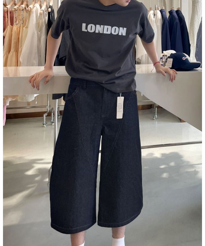 クロップドバレルレッグデニムパンツにグレーのロゴTシャツを合わせ、アパレル店内のカウンターに寄りかかる男性のスタイリング姿。