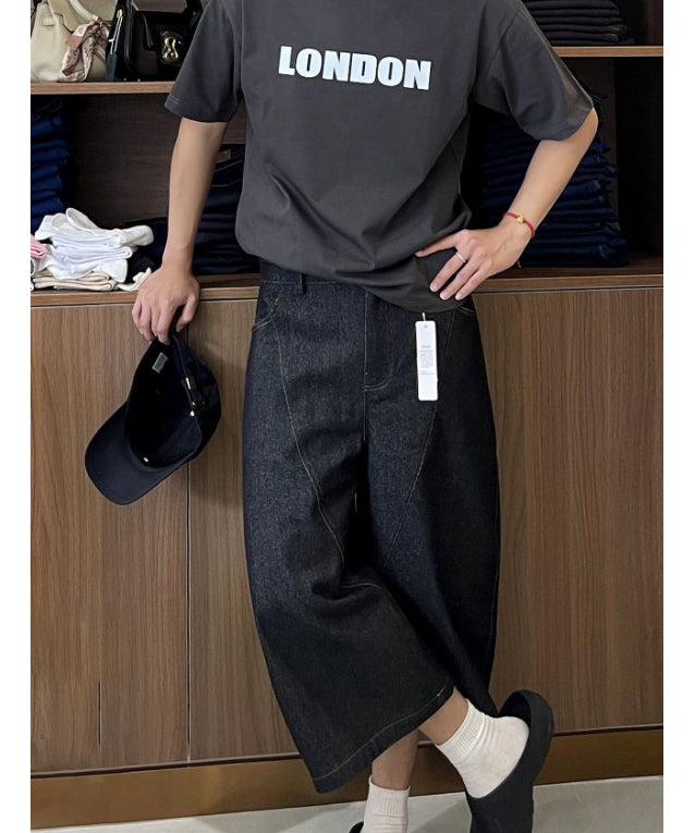 クロップドバレルレッグデニムパンツを着用し、片手に黒いキャップを持って木製棚の前に立つ男性の全身コーディネート。