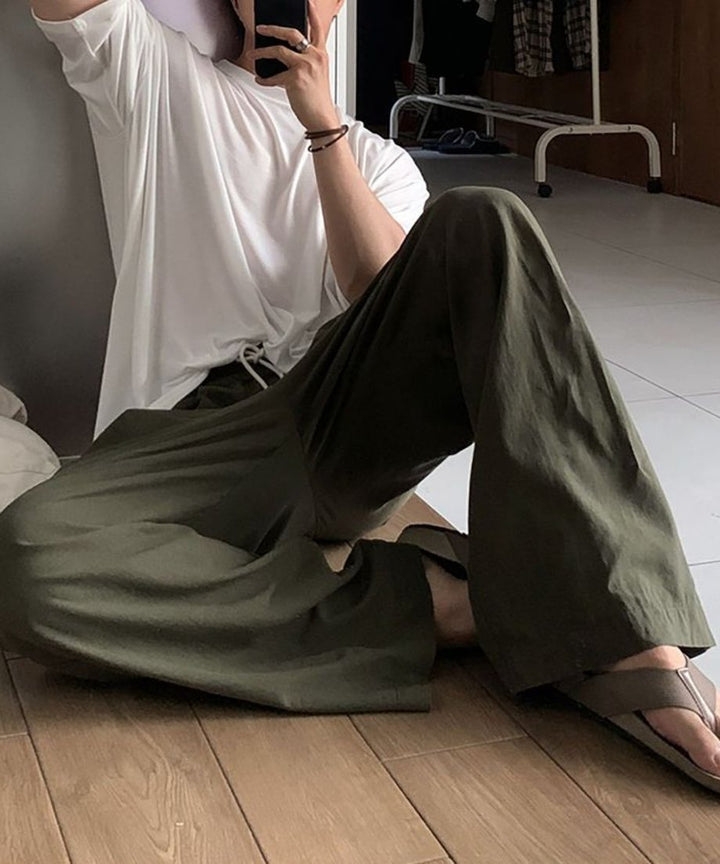 床にリラックスして座り、ホワイトのTシャツとカーキのワイドイージーパンツ、トングサンダルを合わせた男性の自撮りポーズ。