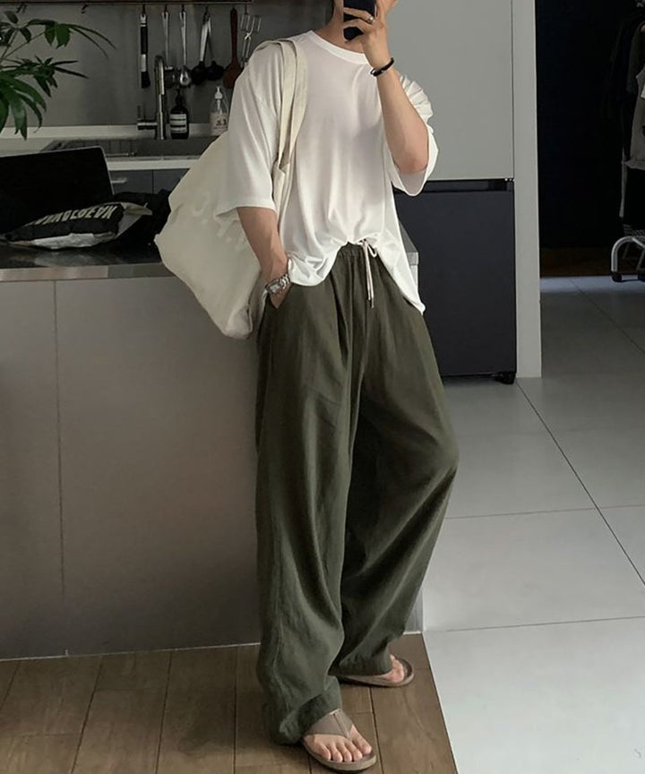 白のオーバーサイズTシャツにカーキのパンツを合わせ、大きめの白いキャンバストートバッグを肩にかけた男性のカフェ風リラックススタイル。