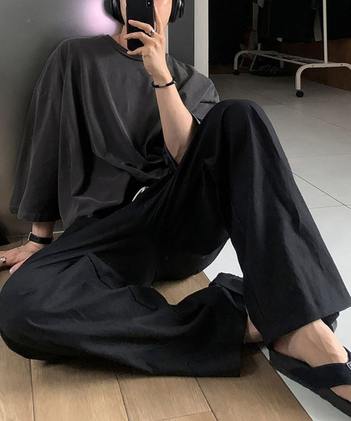 床に座り、壁にもたれかかりながら、アイアングレーの5分袖Tシャツとワイドパンツ、黒のサンダルを合わせたリラクシーな全身スタイリング。