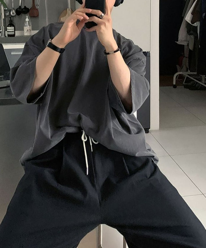 スマホを両手で持って自撮りをする、アイアングレーTシャツと黒パンツを合わせた男性の、ドロップショルダーの落ち感が際立つポーズ。