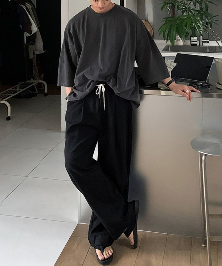 キッチンカウンターに寄りかかり、アイアングレーの5分袖Tシャツと黒のワイドパンツ、トングサンダルを合わせた男性のスタイリッシュな全身スタイリング。