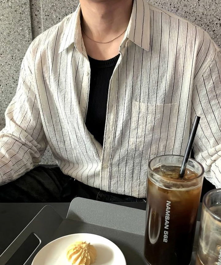 カフェでアイスコーヒーを前に、シャツをタックインして襟元を少し広げ、シルバーのチェーンネックレスを覗かせた男性の着こなし。